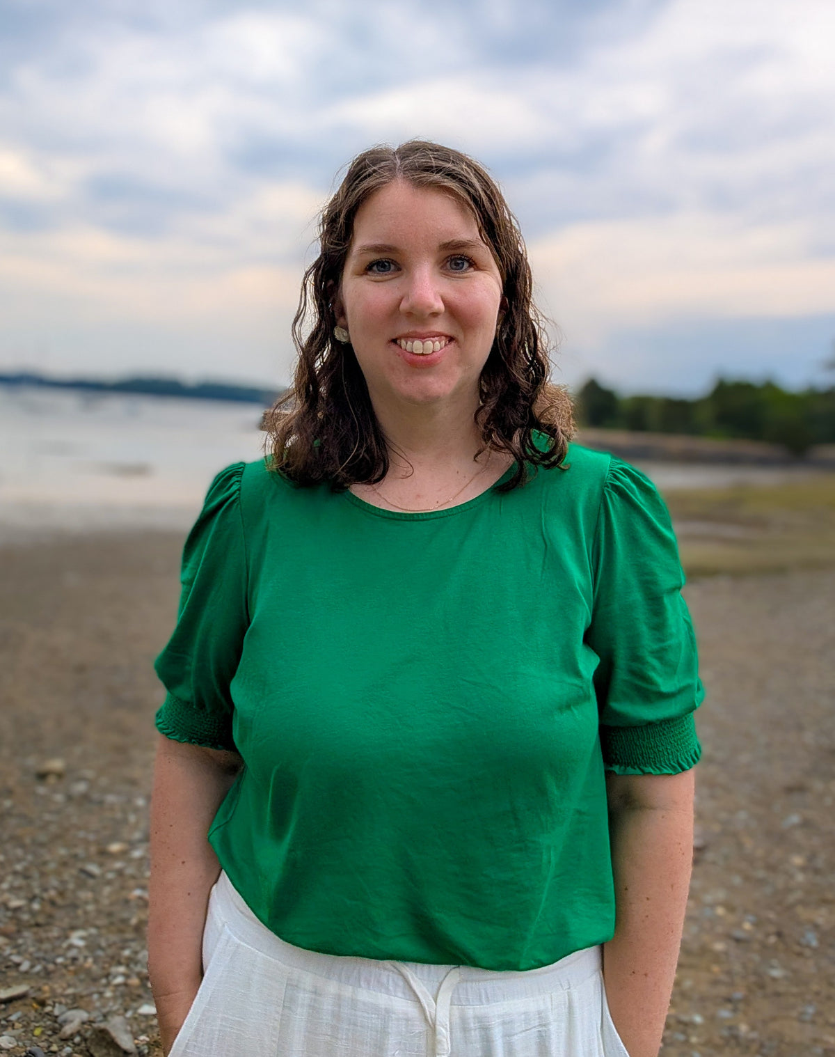 Kaitlyn Nolan, a woman with shoulder-length brown hair stands outdoors on a pebble beach wearing a bright green short-sleeve top and white bottoms, with water and a cloudy sky in the background.